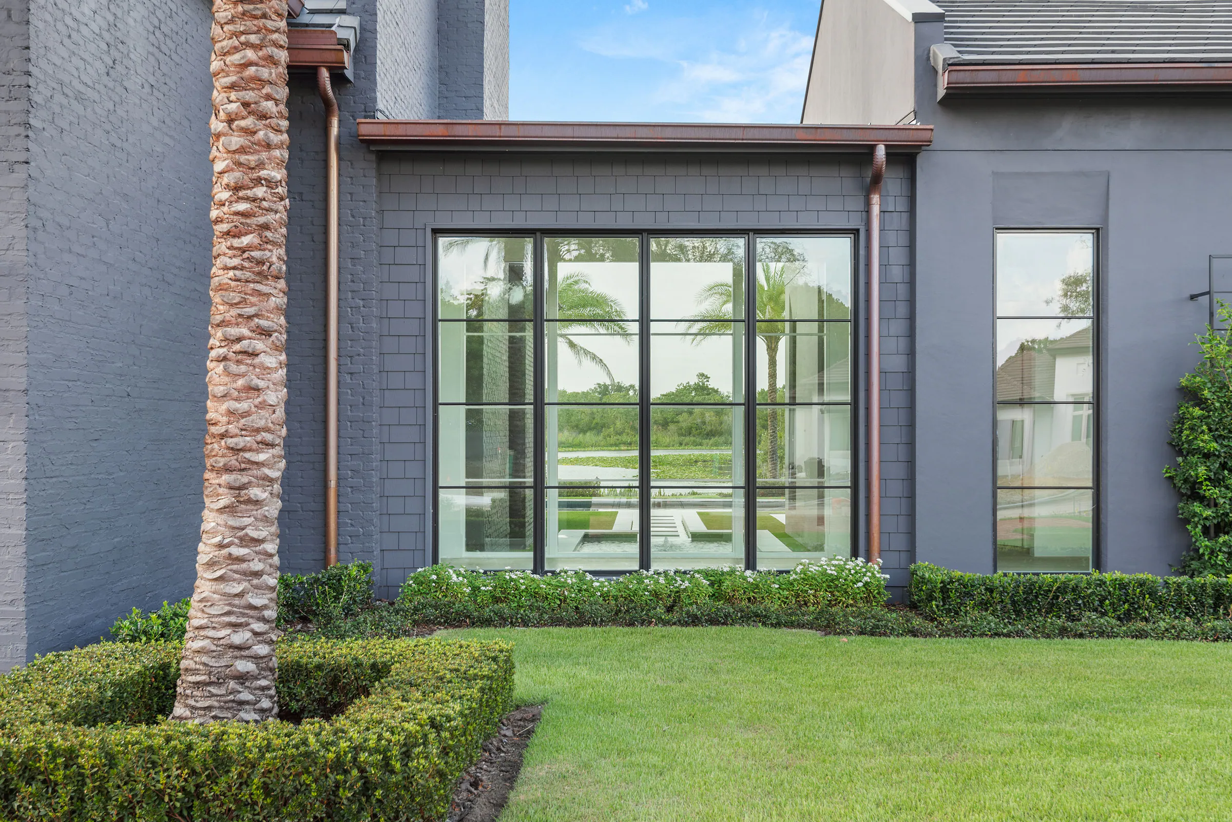 Close view of a black-framed corner window set into dark brick beside a palm and clipped hedges.