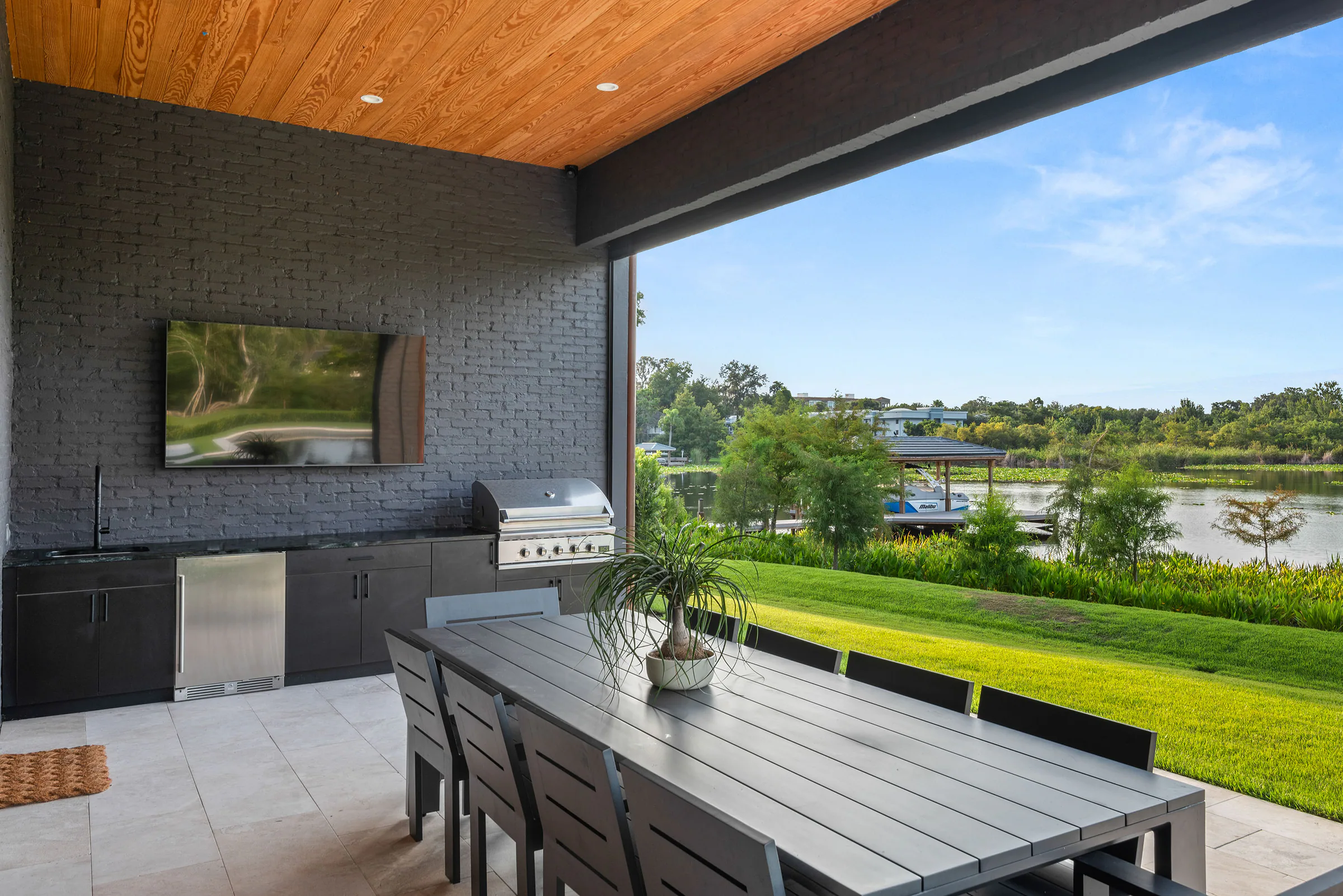 Covered outdoor kitchen with dark brick, wood ceiling, dining table and a view toward the lake.