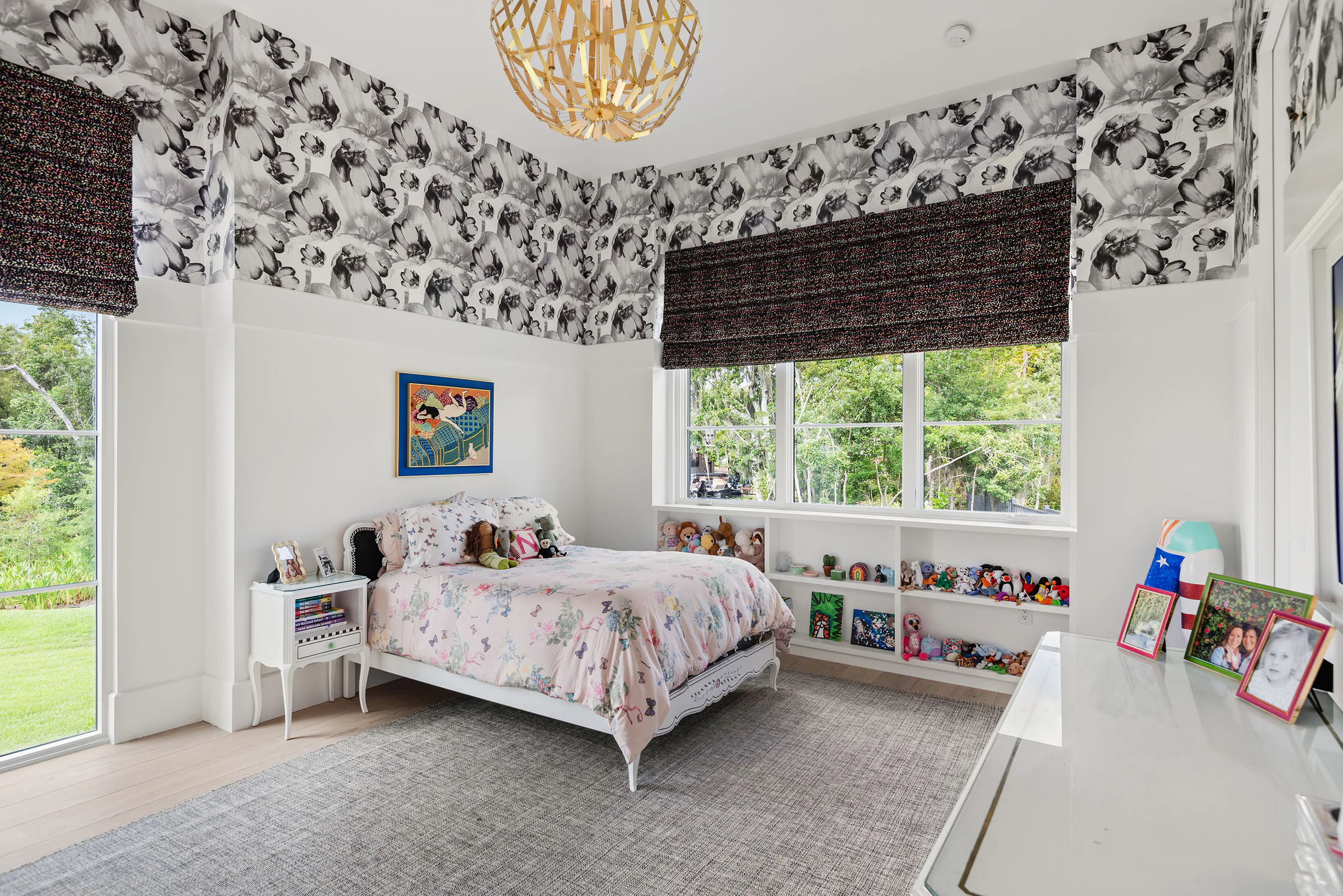 Light-filled bedroom with patterned wallpaper, woven shades and a small white bed between corner windows.