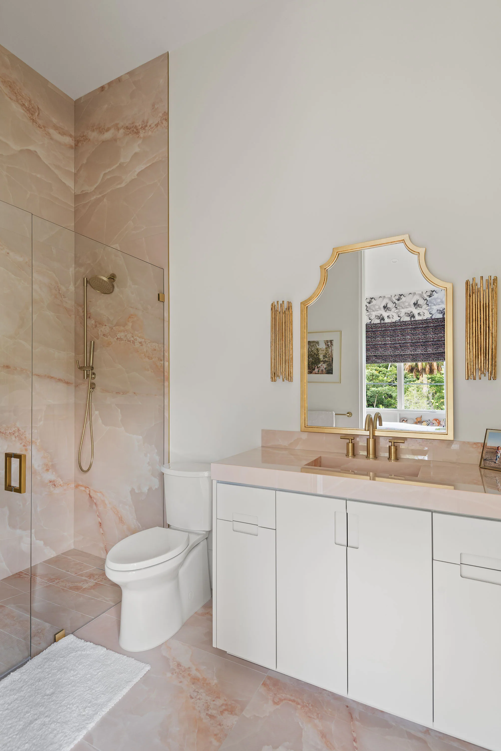 Vertical view of a pale stone bathroom with a floating white vanity, brass fittings and glass shower.