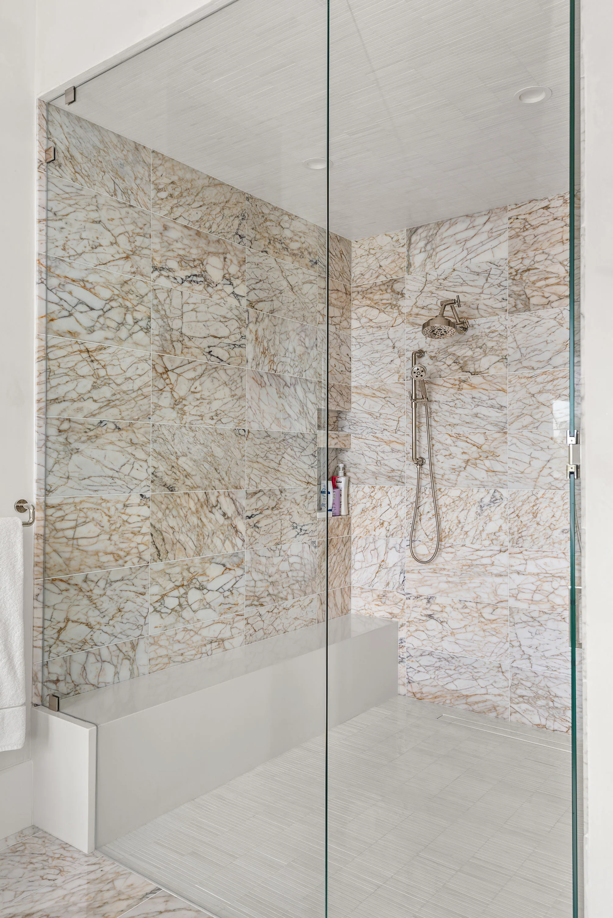 Vertical detail of a glass shower lined with pale mosaic stone tile.