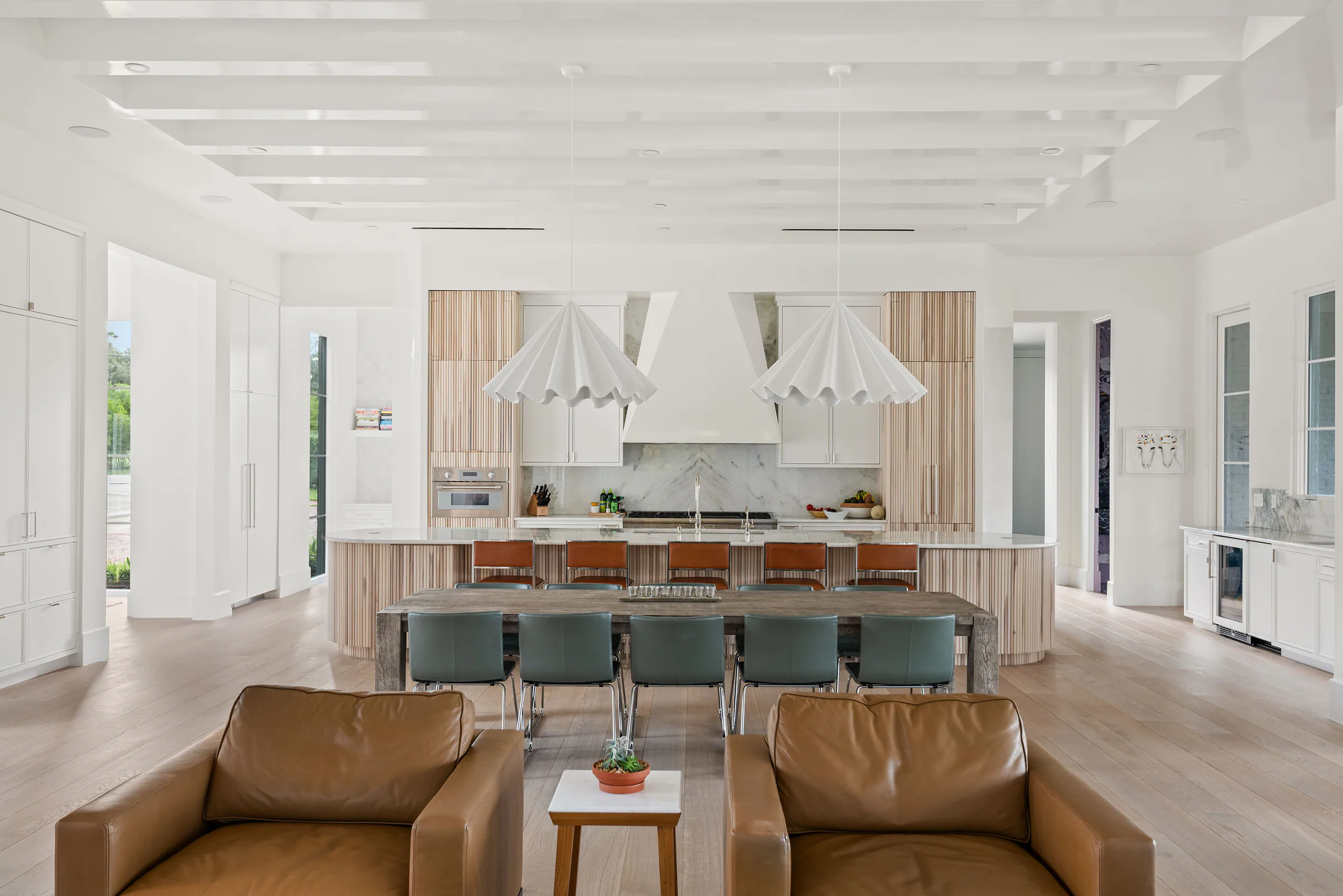 Wide kitchen view with a curved pale wood island, white cabinetry and a long dining table beneath pendant lights.