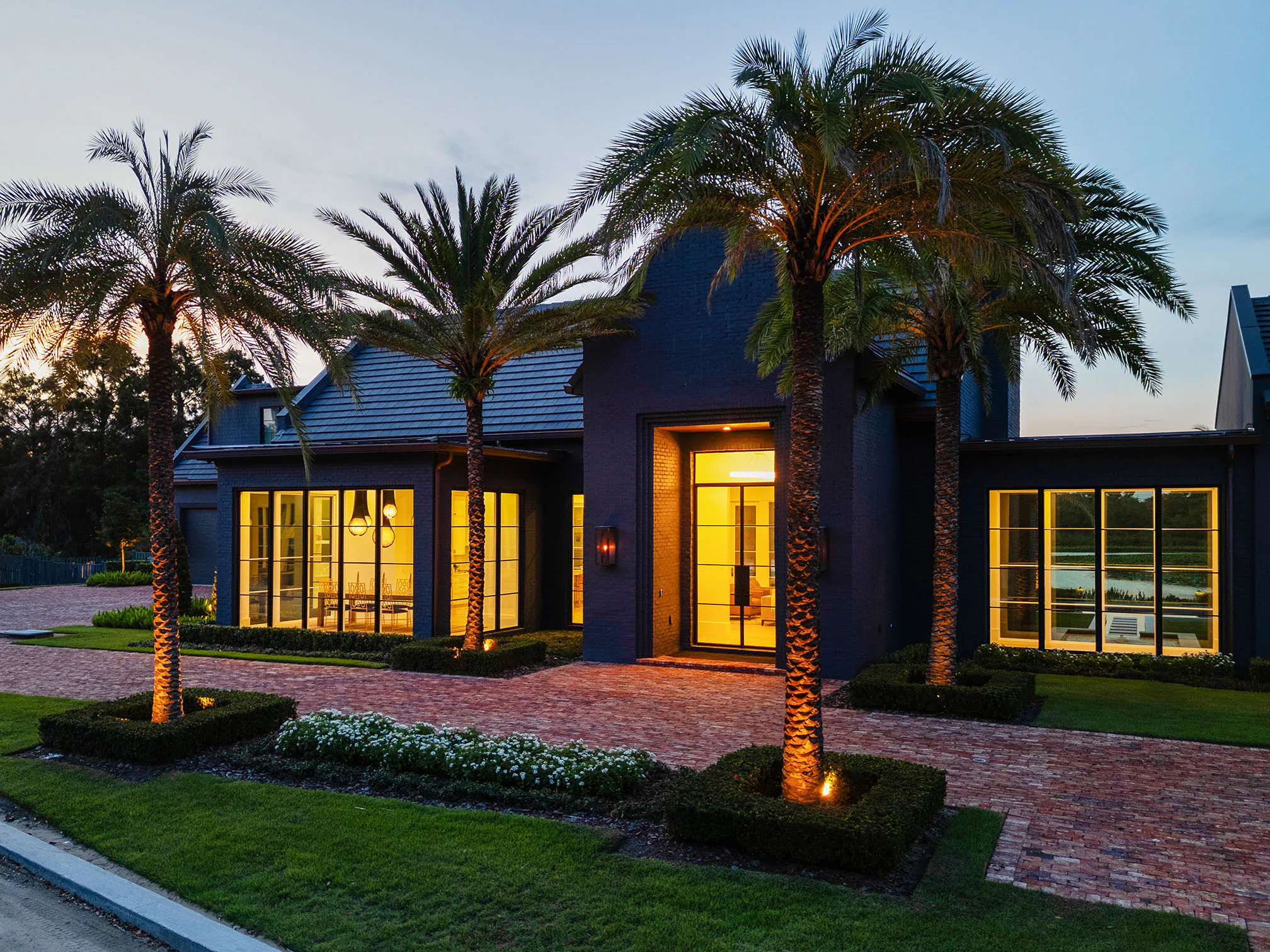 Illuminated front entry of Manor at dusk, framed by palms, clipped hedges and the brick drive.